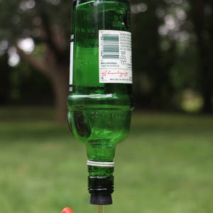 May include: A repurposed green glass bottle bird feeder. The bottle is suspended by a metal hook and ring. A red and clear plastic feeding tube extends from the bottle's bottom. The background is blurred green foliage.