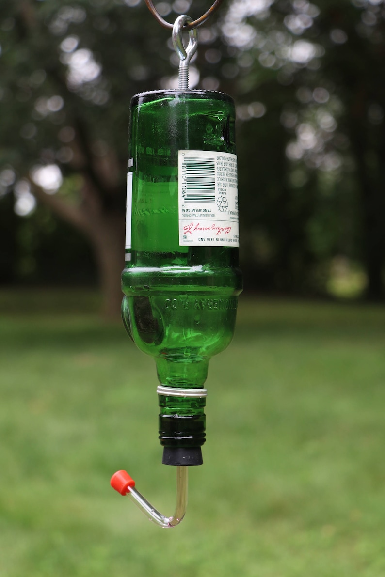 May include: A repurposed green glass bottle bird feeder. The bottle is suspended by a metal hook and ring. A red and clear plastic feeding tube extends from the bottle's bottom. The background is blurred green foliage.