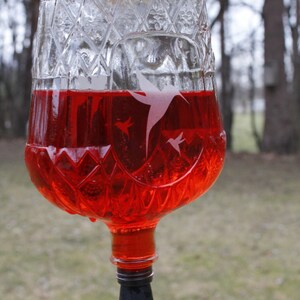 May include: A clear glass hummingbird feeder with a diamond-patterned texture filled with bright red liquid. The glass features etched silhouettes of hummingbirds. A black stopper is at the base with a curved feeding tube and a red tip.