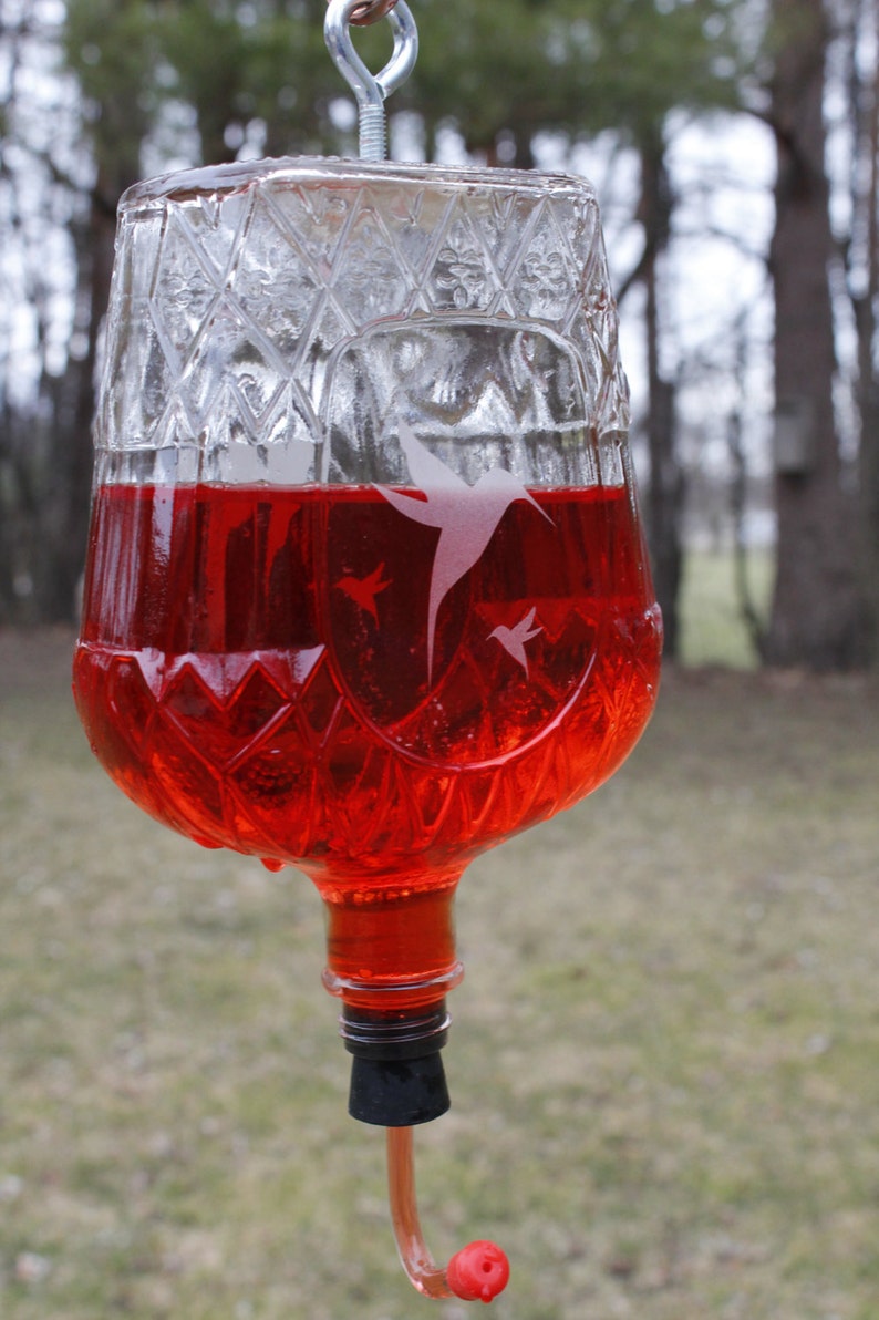 May include: A clear glass hummingbird feeder with a diamond-patterned texture filled with bright red liquid. The glass features etched silhouettes of hummingbirds. A black stopper is at the base with a curved feeding tube and a red tip.