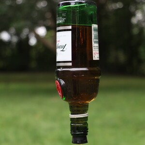 May include: A repurposed glass bottle bird feeder, suspended by a metal hook. The bottle is filled with liquid and has a red and clear feeding tube attached. The bottle has a green top and a white label with black text.