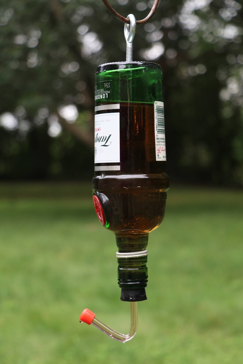 May include: A repurposed glass bottle bird feeder, suspended by a metal hook. The bottle is filled with liquid and has a red and clear feeding tube attached. The bottle has a green top and a white label with black text.