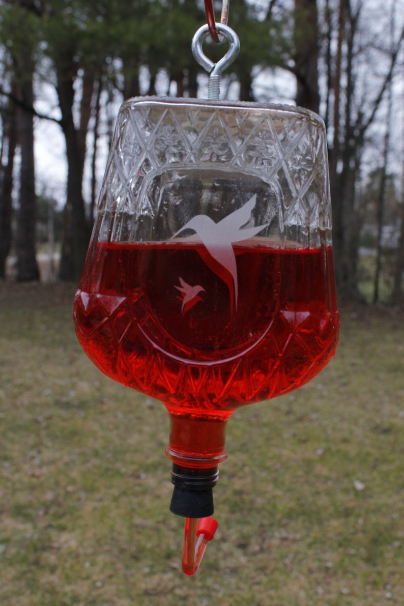 May include: A clear glass hummingbird feeder with a diamond-cut pattern filled with red liquid. The feeder features a white etched graphic of two hummingbirds and is suspended by a silver metal eye bolt attached to the base of the glass vessel.
