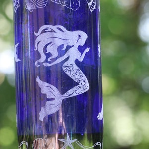 May include: A blue glass wind chime with a mermaid design. The wind chime is made from a repurposed bottle and features etched seashells and a starfish. The mermaid is etched in white and has flowing hair and a decorative tail.