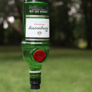 May include: A repurposed green glass bottle, labeled "Tanqueray London Dry Gin," transformed into a bird feeder. The bottle is suspended by a metal hook, with a red stopper and a clear tube with a red ball at the end for dispensing birdseed.