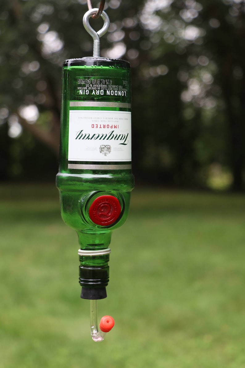 May include: A repurposed green glass bottle, labeled "Tanqueray London Dry Gin," transformed into a bird feeder. The bottle is suspended by a metal hook, with a red stopper and a clear tube with a red ball at the end for dispensing birdseed.