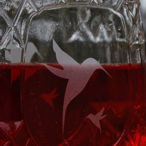 May include: A clear glass vessel with a diamond-cut textured pattern filled with a vibrant red liquid. Etched onto the surface of the glass are three hummingbird silhouettes of varying sizes, appearing frosted against the transparent glass.