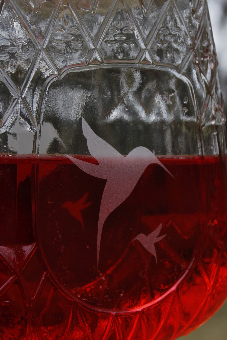 May include: A clear glass vessel with a diamond-cut textured pattern filled with a vibrant red liquid. Etched onto the surface of the glass are three hummingbird silhouettes of varying sizes, appearing frosted against the transparent glass.