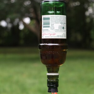 May include: A repurposed green glass bottle, suspended by a metal hook, with a clear liquid dripping from the bottom. The bottle contains a brown liquid and has a label with text. The background is blurred green foliage.