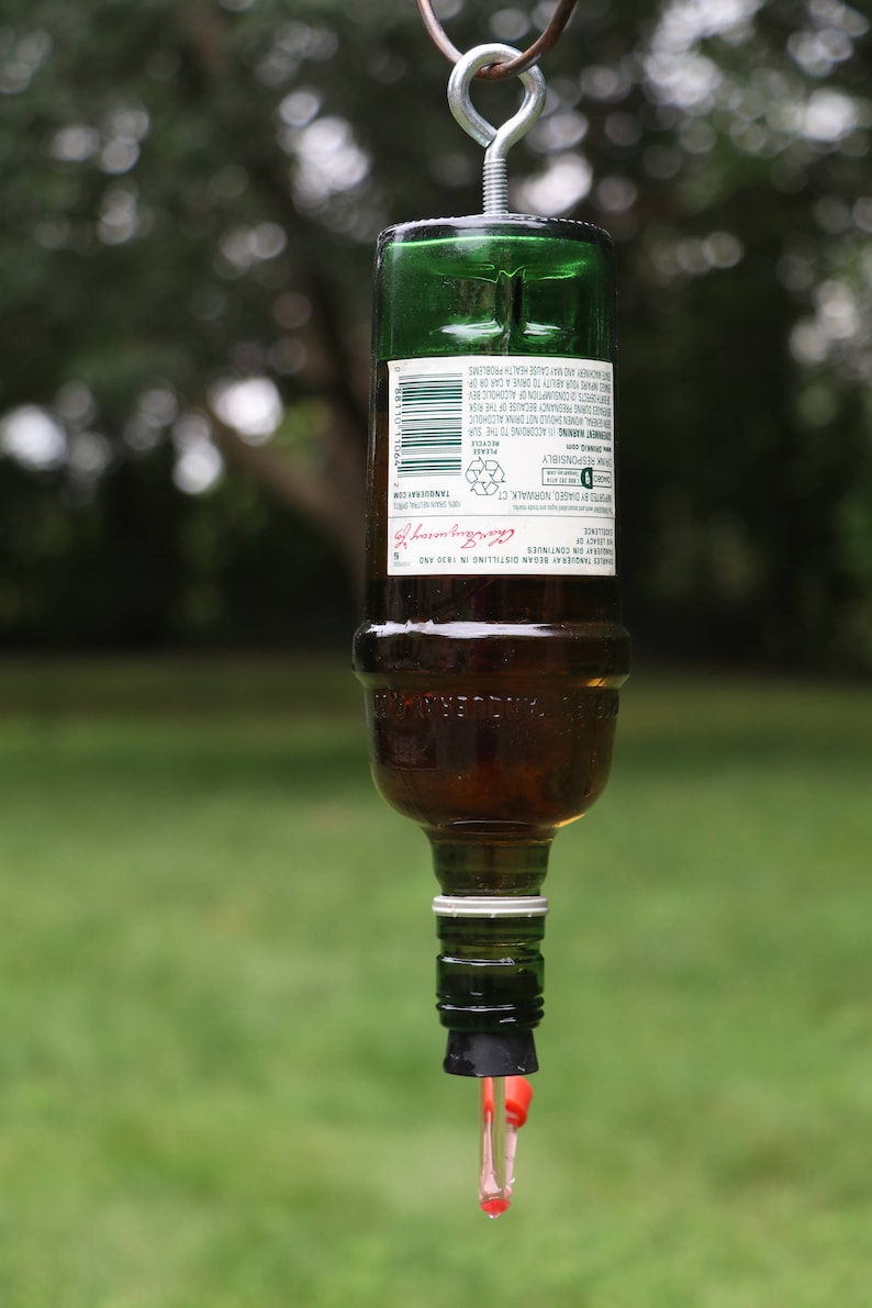 May include: A repurposed green glass bottle, suspended by a metal hook, with a clear liquid dripping from the bottom. The bottle contains a brown liquid and has a label with text. The background is blurred green foliage.