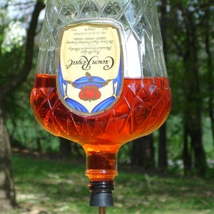Op de afbeelding: Een omgekeerde Crown Royal glazen fles gevuld met oranje vloeistof, met een ruitvormige textuur en een etiket met een kroonafbeelding. Een oogbout is aan de basis bevestigd, met een zwarte stop met een gebogen tuit en een oranje punt.