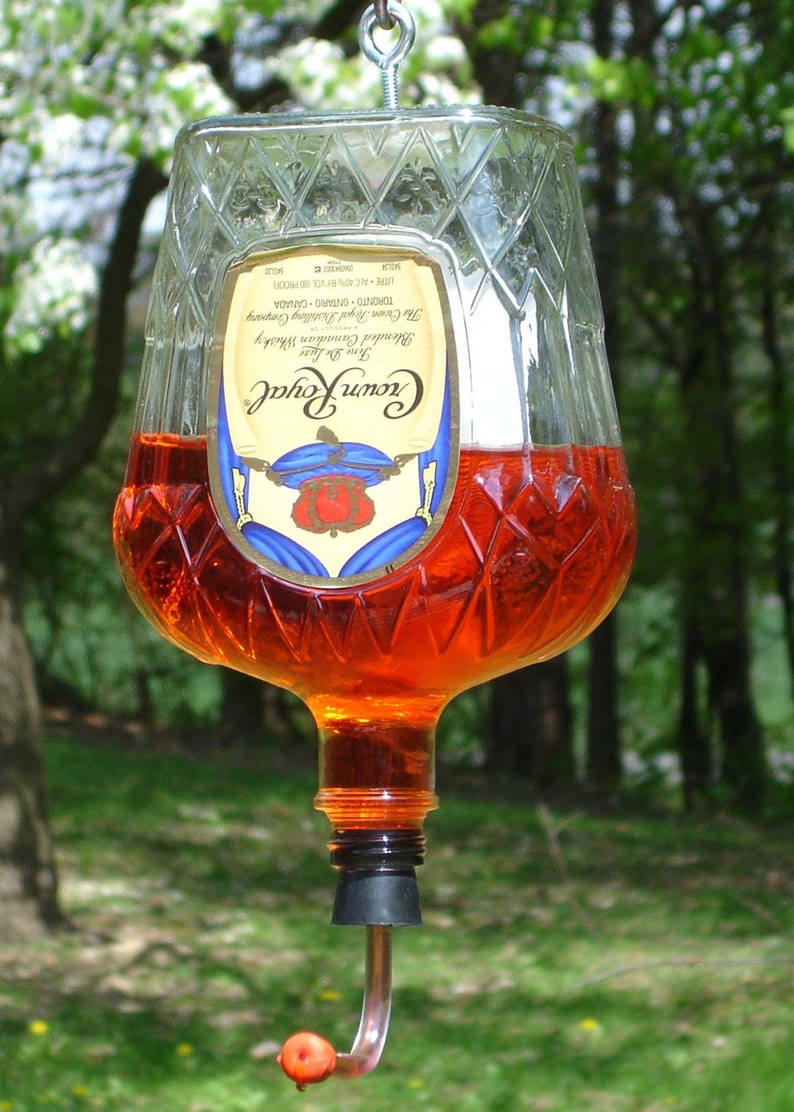 Op de afbeelding: Een omgekeerde Crown Royal glazen fles gevuld met oranje vloeistof, met een ruitvormige textuur en een etiket met een kroonafbeelding. Een oogbout is aan de basis bevestigd, met een zwarte stop met een gebogen tuit en een oranje punt.