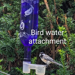 Op de afbeelding: Een kobaltblauwe glazen fles met witte gegraveerde zeemeermin- en schelpmotieven, voorzien van een doorzichtig plastic vogeldrinkbakje aan de onderkant. Een kleine vogel met zwarte, witte en grijze veren zit op de rand van het bakje.