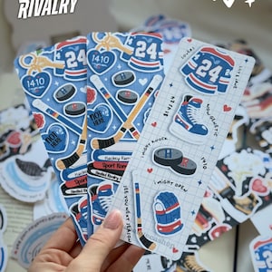 Könnte beinhalten: Eine Hand hält mehrere Hockey-Sticker in Blau, Rot und Weiß. Die Sticker zeigen Hockey-Trikots, Schlittschuhe, Pucks und Schläger. Text enthält "HEATED RIVALRY", "24" und "1410".
