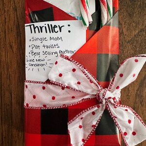May include: A red and black plaid gift box with a white bow and red polka dots. The box is wrapped with a white paper note that says "Thriller: Single MOM Plot twists Best Selling Author Like New Condition!" and a candy cane is tucked inside the box.