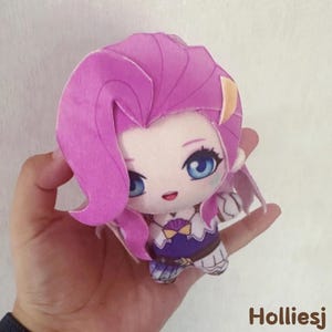 May include: A plush doll with a cartoon-style design. The doll has large blue eyes, pink hair, and a detailed outfit with a blue top and white accents. The doll is held in a hand. The text "Holliesj" is in the bottom right corner.