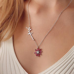 May include: A silver necklace with a flower pendant featuring pink gemstones and a central clear stone. A matching bracelet with a flower design and blue gemstones is also visible. Both pieces have the name "Meghan" inscribed.