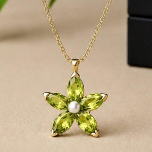 May include: A gold chain necklace featuring a flower-shaped pendant. The pendant consists of five marquise-cut lime green gemstones arranged as petals around a central small white pearl. The necklace is suspended from a thin gold-toned metal chain.