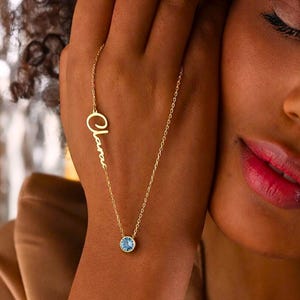 May include: A gold-colored necklace with the name "Clara" in cursive script and a round, light blue gemstone pendant. The necklace is displayed on a person's skin, with a focus on the jewelry's delicate design and color contrast.