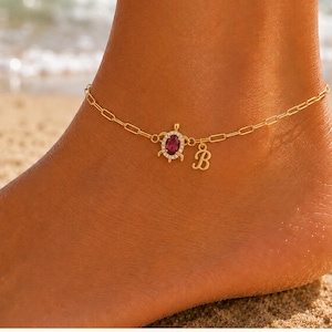 May include: Gold-toned anklet with a turtle charm featuring a red gemstone and a letter "B" pendant. The anklet is worn on a tanned ankle, with the ocean in the background.
