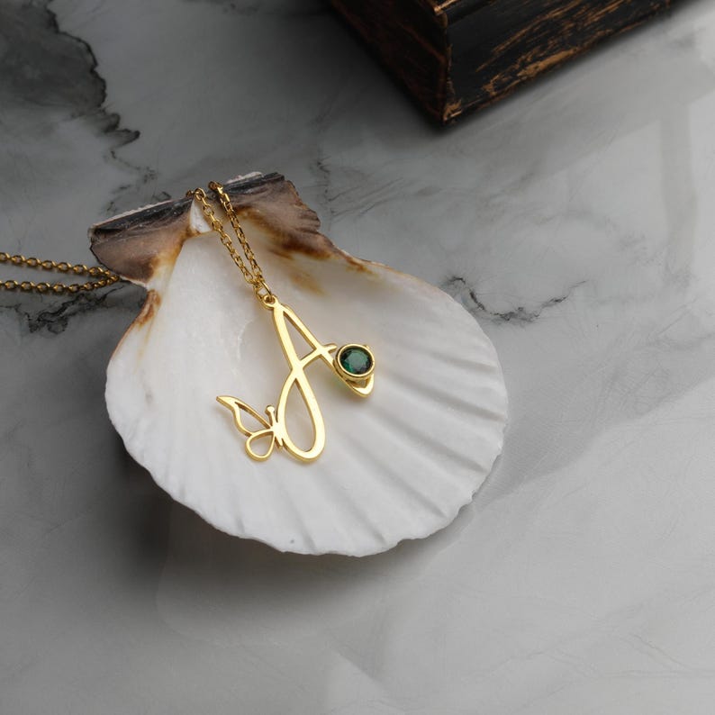 May include: A gold-toned necklace featuring a pendant shaped like the letter A with a small butterfly detail at the base and a single round, dark green gemstone set into the upper right curve of the letter, suspended from a delicate gold chain.