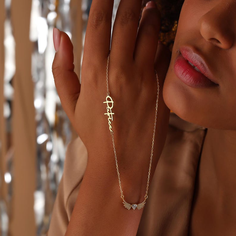 Pu&ograve; includere: Una collana dorata con il nome "Patrice" in corsivo. Il ciondolo presenta ali d'angelo e una piccola gemma trasparente. La collana &egrave; mostrata sulla mano di una persona. Lo sfondo &egrave; sfocato.