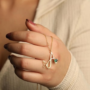 May include: A gold-toned necklace featuring a cursive letter A pendant. The pendant is accented with a small butterfly outline on the left side and a round, faceted emerald green gemstone set on the right side of the letter, suspended on a chain.