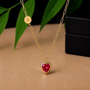 May include: A gold-toned chain necklace featuring a heart-shaped red gemstone pendant and a small circular charm engraved with the letter A. The chain consists of elongated rectangular links, and the pendant is set in a smooth gold-toned metal frame.