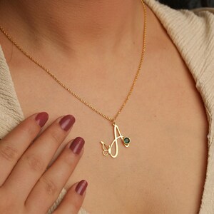 May include: A gold-toned necklace featuring a cursive letter A pendant integrated with a small butterfly outline and a single round dark green gemstone. The pendant hangs from a delicate gold-toned chain resting against a textured beige fabric.