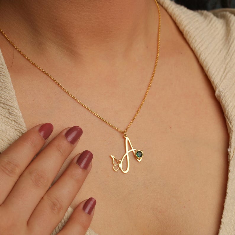 May include: A gold-toned necklace featuring a cursive letter A pendant integrated with a small butterfly outline and a single round dark green gemstone. The pendant hangs from a delicate gold-toned chain resting against a textured beige fabric.