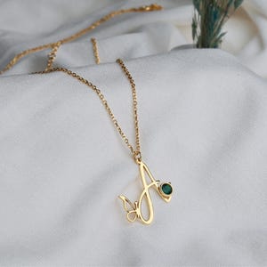 May include: A gold-toned necklace featuring a cursive letter A pendant. The pendant is accented with a small butterfly silhouette on the left side and a single round, dark green gemstone set into the upper right curve of the letter.