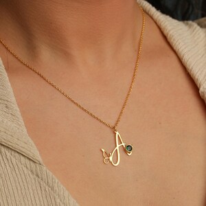 May include: A gold-toned necklace featuring a cursive letter A pendant. The pendant is accented with a small butterfly silhouette on the left side and a single round, dark green gemstone set into the right side of the letter's stroke.