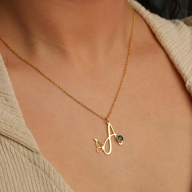 May include: A gold-toned necklace featuring a cursive letter A pendant. The pendant is accented with a small butterfly silhouette on the left side and a single round, dark green gemstone set into the right side of the letter's stroke.