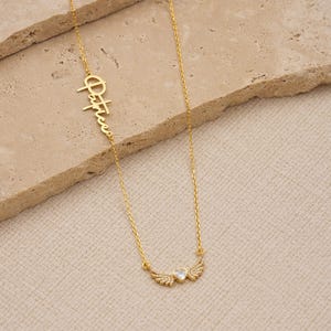 Pu&ograve; includere: Una collana dorata con una catena delicata. La collana presenta il nome "Patrice" in corsivo, insieme a un ciondolo a forma di cuore con ali di cristallo. La collana &egrave; esposta su una superficie strutturata di colore chiaro.