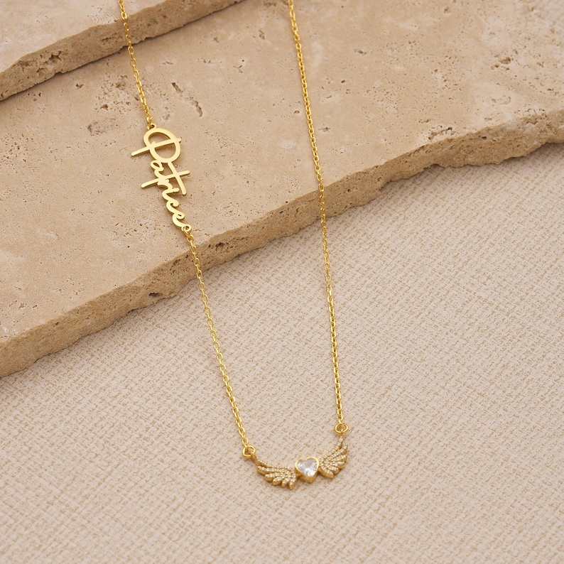 Pu&ograve; includere: Una collana dorata con una catena delicata. La collana presenta il nome "Patrice" in corsivo, insieme a un ciondolo a forma di cuore con ali di cristallo. La collana &egrave; esposta su una superficie strutturata di colore chiaro.