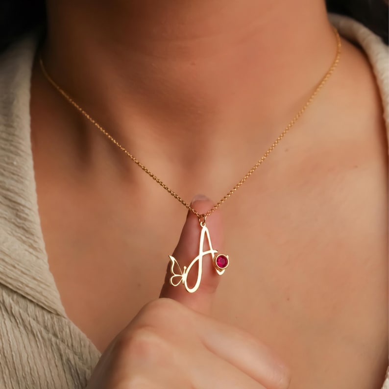 May include: A gold-toned necklace featuring a cursive letter A pendant. The pendant is accented with a small, delicate butterfly silhouette on the left side and a single, round, vibrant red gemstone set on the right side of the letter.