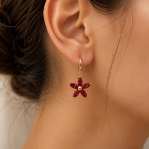 May include: A gold hoop earring featuring a dangling flower charm. The flower is composed of five marquise-cut deep red gemstones arranged around a small central gold bead, creating a floral silhouette that hangs from a simple gold hoop.