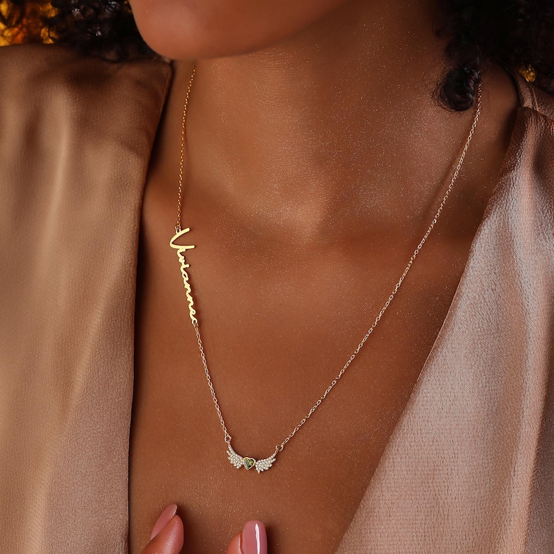 Pu&ograve; includere: Una collana color oro con il nome "Vivianne" in corsivo. La collana presenta un ciondolo con dettagli a forma di ali e una piccola pietra preziosa verde. La collana &egrave; indossata intorno al collo.