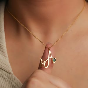 May include: A gold-toned necklace featuring a cursive letter A pendant. The pendant is adorned with a small butterfly cutout on the left side and a single round, dark green gemstone set into the right side of the letter. The chain is thin and metallic.