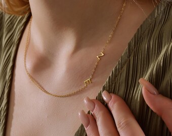 May include: A gold-colored necklace with the letters "E" and "N" is displayed. The necklace is worn around the neck. The background is a green-colored top. The necklace is a delicate piece of jewelry.