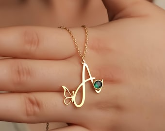 May include: A gold-toned necklace featuring a cursive letter A pendant. The pendant is accented with a small butterfly silhouette on the left side and a single round, dark green gemstone set in a gold-toned bezel on the right side.