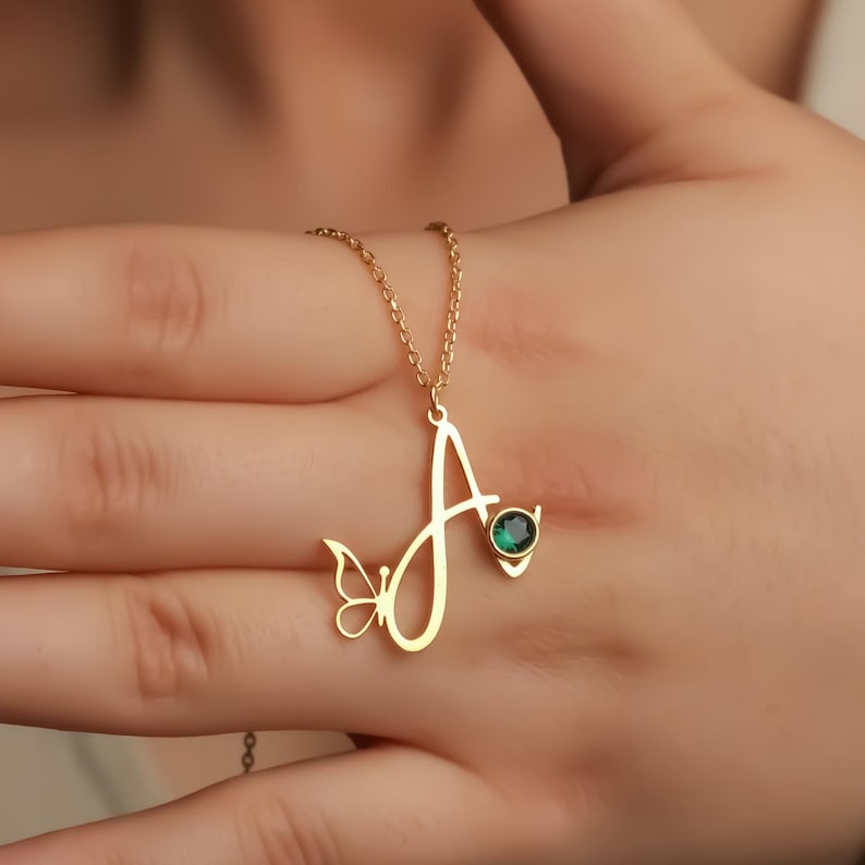 May include: A gold-toned necklace featuring a cursive letter A pendant. The pendant is accented with a small butterfly silhouette on the left side and a single round, dark green gemstone set into the right side of the letter, suspended on a chain.