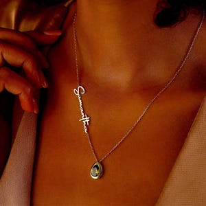May include: A delicate silver necklace with the name "Samantha" in cursive script. The necklace features a teardrop-shaped pendant with a dark center and yellow accents. The necklace is worn on a person's neck.