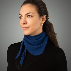 Puede incluir: Una bufanda azul se lleva alrededor del cuello. La bufanda es de un tono azul profundo y está atada con un nudo en la parte delantera. La bufanda está hecha de un material suave. La persona lleva una camisa negra de manga larga.