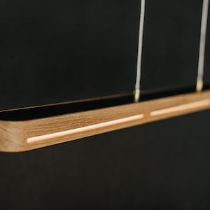 Könnte beinhalten: Eine moderne Holz-Pendelleuchte mit schwarzem Innenraum und zwei integrierten LED-Streifen. Die Leuchte wird mit zwei dünnen Schnüren von der Decke abgehängt.