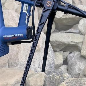 Op de afbeelding: Een blauwe en zwarte metaaldetector met de tekst "GOLD MONSTER 1000". De detector heeft een zwarte handgreep en een ondersteunende structuur. De afbeelding is tegen een achtergrond van grijze rotsen geplaatst.