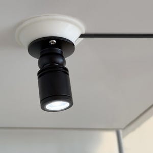 May include: A black cylindrical spotlight fixture mounted to a white surface with a white circular base. The light features a swivel joint for directional adjustment and emits a bright white glow from the circular lens at the bottom of the unit.