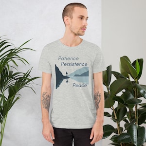 Puede incluir: Una camiseta gris claro con un gráfico de una persona en una montaña, con las palabras "Paciencia, Persistencia, Paz" en azul. El diseño incluye un reflejo en el agua. La camiseta está hecha de un material suave.