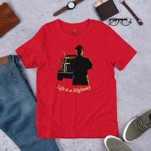 Puede incluir: Una camiseta roja con un gráfico de un camión y una persona con los brazos cruzados. El texto "Life is a Highway" está impreso en cursiva debajo del gráfico. La camiseta está hecha de un material suave.