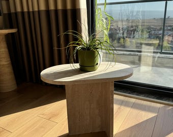 May include: A small, oval-shaped, light beige travertine side table with a single, thick, angled leg on each side. The table is sitting on a light wood floor and has a small green potted plant on top.